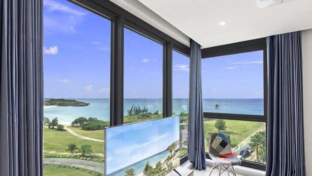 A modern living room with a large TV on a stand, a comfortable chair, and a view of a beach and ocean through large windows. - A601 (Cupecoy)