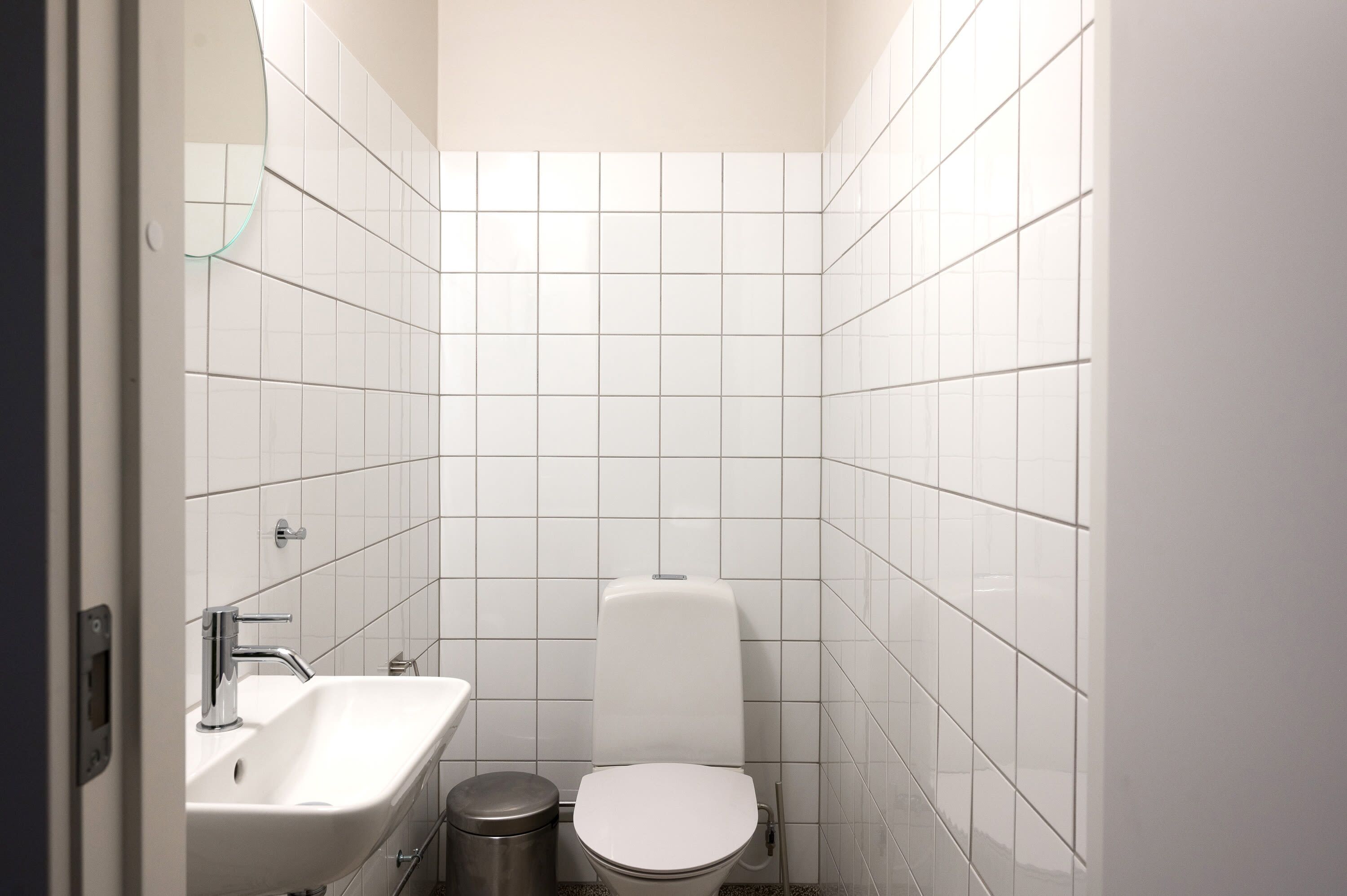 Et lille badeværelse med hvide fliser, et håndvask, et toilet og en skraldespand.