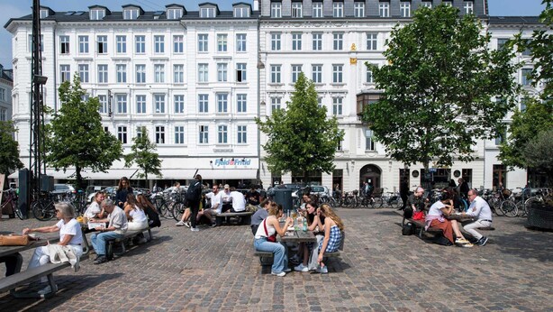 Julkinen aukio, jossa ihmiset istuvat pöytien ääressä. Aukiota ympäröivät historialliset rakennukset, joissa on suuret ikkunat, ja näkyvillä on kyltti, jossa lukee ”Feld & Fritid”.