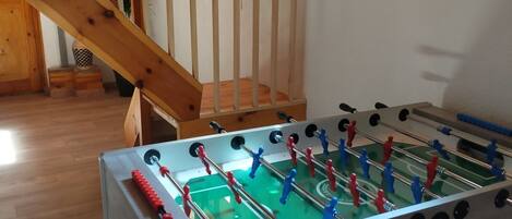 A room with a foosball table, wooden flooring, a staircase, and a window with natural light.