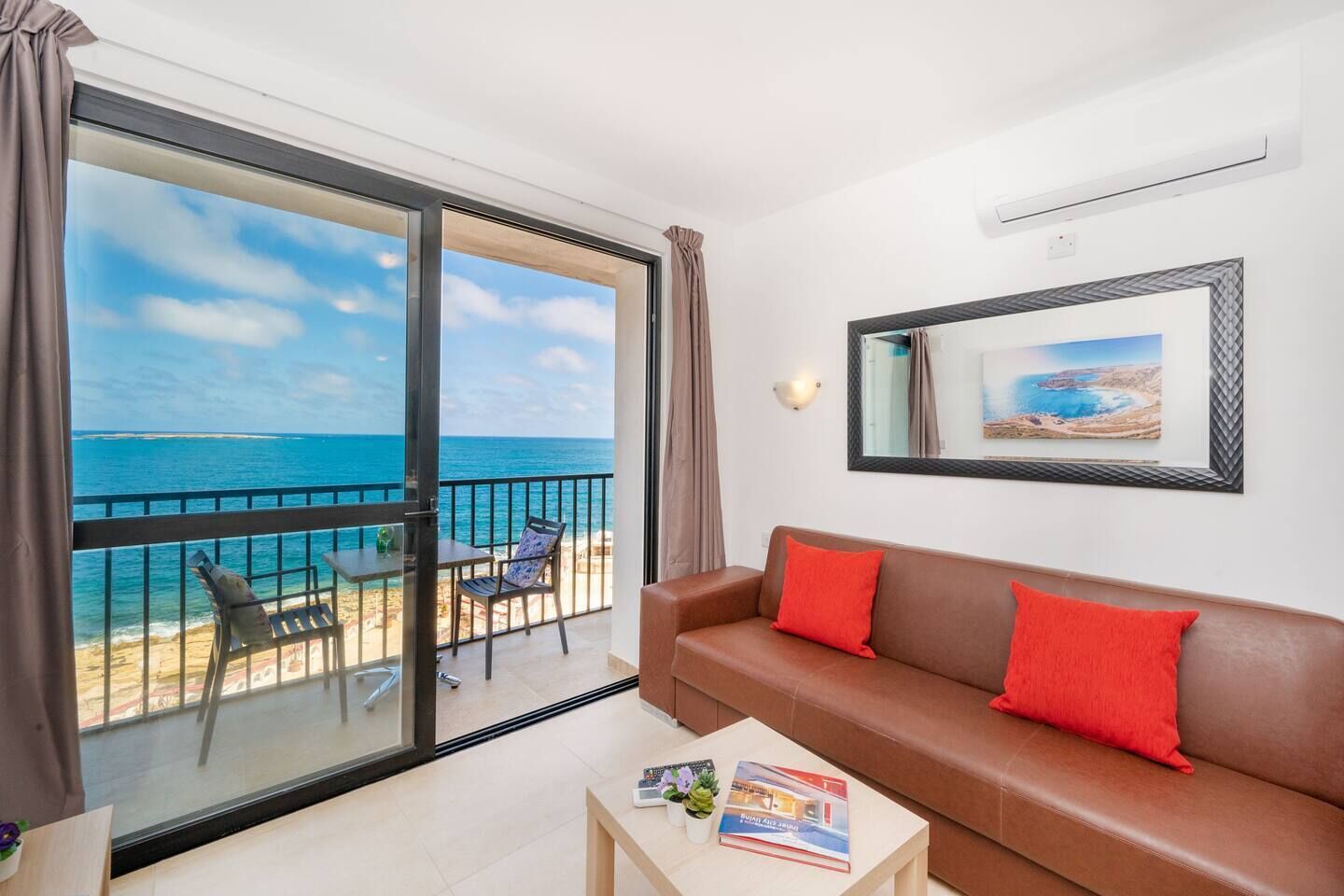A balcony with a view of the sea, a brown leather sofa with red cushions, a small table with a magazine and a plant, and a framed picture on the wall.