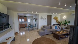 Ein modernes Wohnzimmer mit einem grauen Sofa, einem Couchtisch und einem Fernseher. Im Hintergrund befindet sich eine Küche mit einem Essbereich.