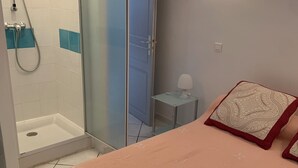 A bedroom with a bed, a nightstand, a lamp, a wall-mounted light, a shower area, and a door. - Appartement 2 en Pierre (Ota)