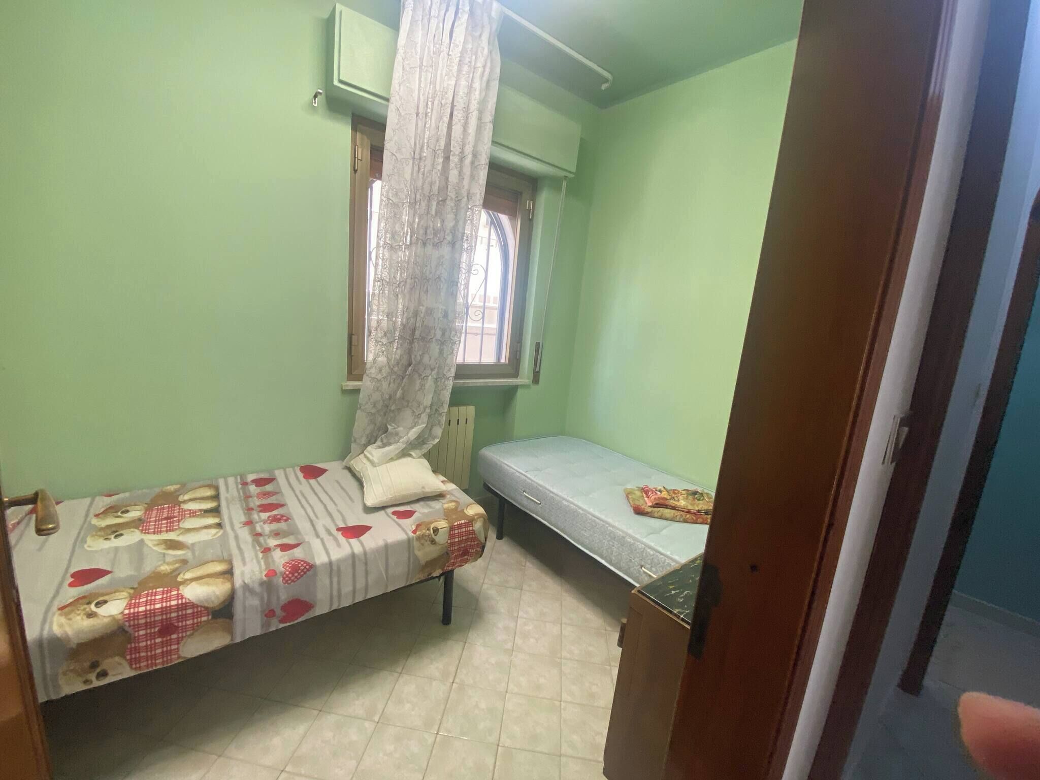 A small, single-room with two beds, a window with a curtain, and a radiator.