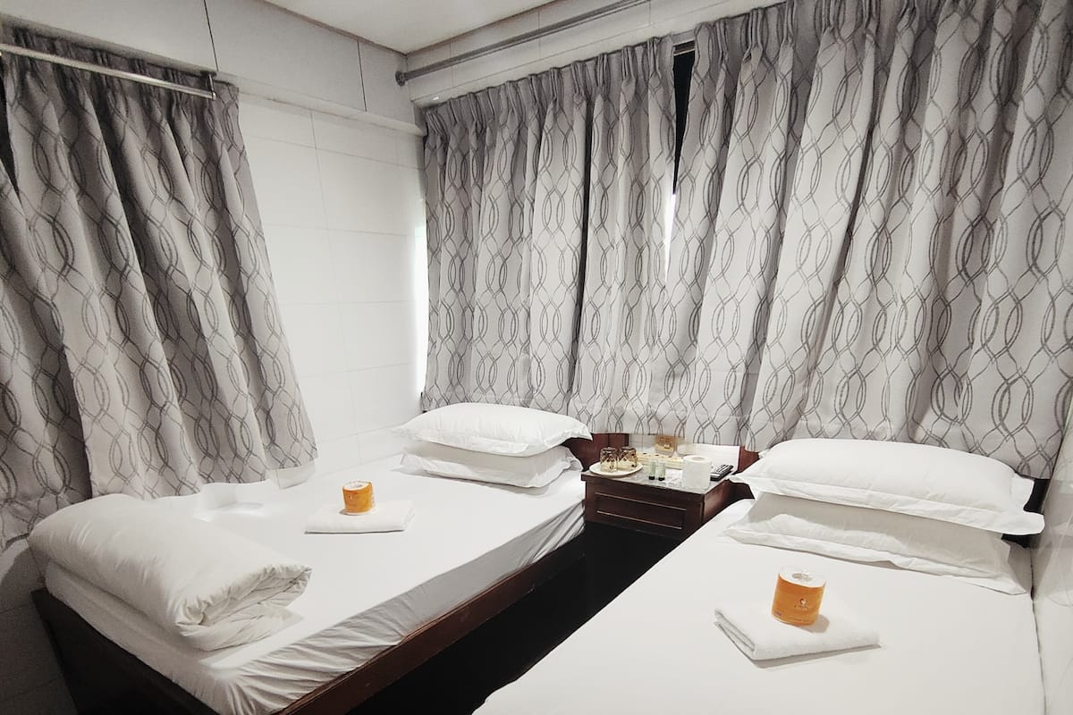 Two single beds with white linens and pillows, a nightstand with a remote, bottle, and tray, and patterned curtains.