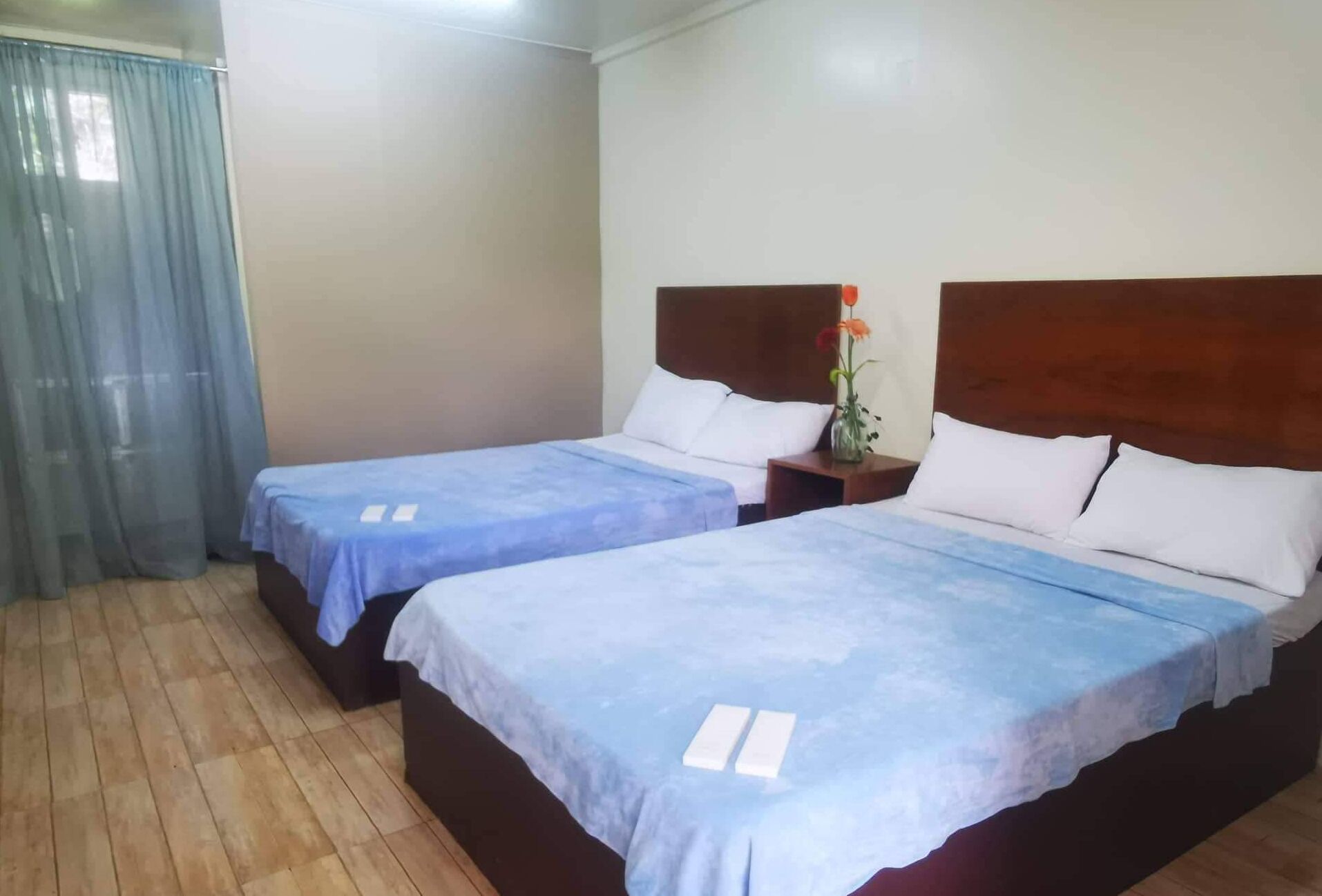 Two single beds with wooden headboards, a nightstand with a vase of flowers, and a window with a sheer curtain.