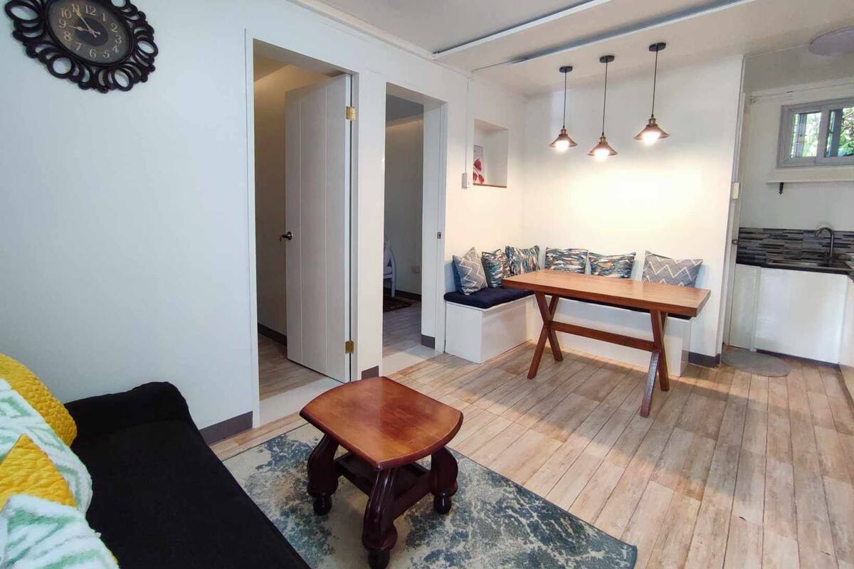 A living room with a sofa, a wooden coffee table, a dining table with benches, and a kitchen area with a sink and countertop.