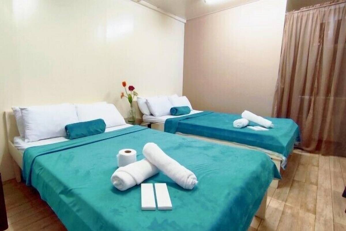 A hotel room with two beds, each with a teal bedspread and white pillows. There are towels and a roll of toilet paper on the beds.