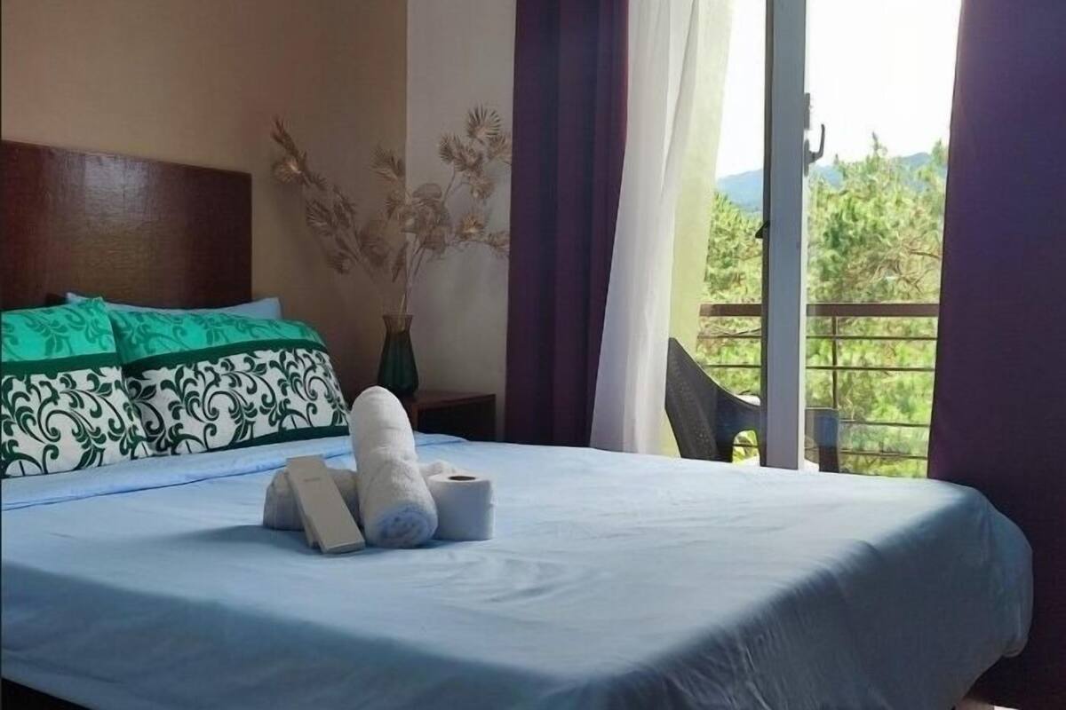 A neatly made bed with a blue bedspread, a green and white patterned pillow, and a vase with dried flowers.