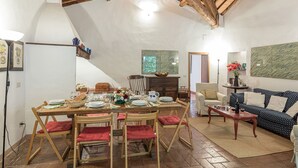 Uma área de refeições com uma mesa de madeira posta para quatro pessoas, rodeada por cadeiras de madeira com almofadas vermelhas. Um armário de madeira rústica e uma pequena mesa lateral com um vaso de flores são visíveis. O quarto apresenta vigas de madeira expostas no teto e um quadro abstrato grande na parede.