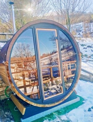 A large, transparent cylindrical structure with a wooden interior, reflecting a snowy landscape and trees. - Maison en Pierre (Fiastra)
