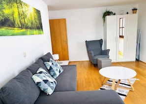 A living room with a sofa, armchair, and a white table. There's a wooden door, a cabinet with plants, and a painting on the wall.