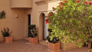 A courtyard with a building featuring a tiled roof and stucco walls. There are potted plants and a small tree with red flowers. - Spectacular mountain views in a romantic location. Close to Mojacar beaches. (Turre)