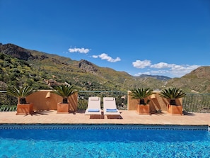 A pool with two sun loungers and potted plants, surrounded by mountains and clear skies.