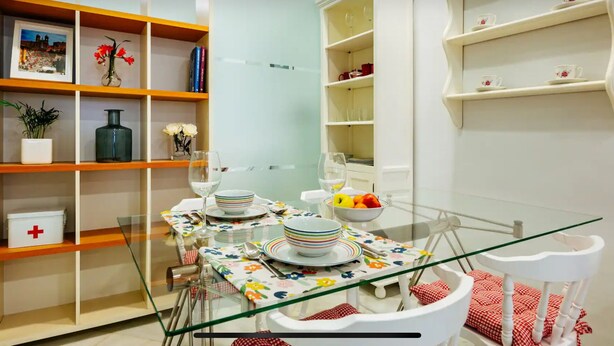 Ein Essbereich mit einem Glastisch, weißen Stühlen und einer Tischdecke mit Blumenmuster. Regale mit Büchern und Dekorationselementen sowie eine weiße Kommode mit Glastüren sind ebenfalls vorhanden.
