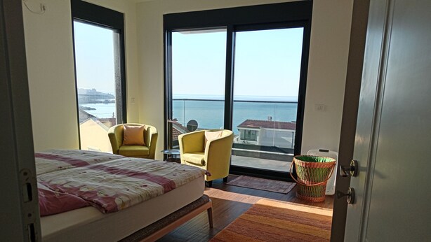 A bedroom with a bed, two yellow chairs, a small table, a wicker basket, and a view of the sea through large windows. - Camp Virpazar (Virpazar)