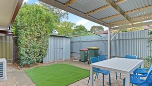 A covered patio area with a table and blue chairs, a green lawn, and a corrugated metal fence.