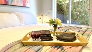 A neatly made bed with a wooden tray holding books and a towel, a vase with flowers, and a window with a view of greenery.