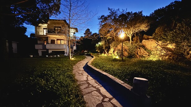 Un sendero de piedra que lleva a una casa de dos pisos con balcón, rodeada de árboles y un jardín bien iluminado.