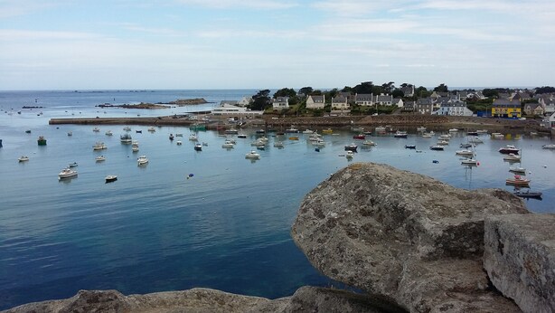 Un pueblo costero con numerosos barcos amarrados en un puerto, rodeado de edificios residenciales y terreno rocoso.