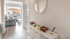 A modern bedroom with a grey sofa, a small table, a dresser with drawers, a mirror, and a view of mountains through a window.