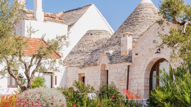 Un edificio de piedra tradicional con tejado de tejas, rodeado de plantas en flor y árboles.