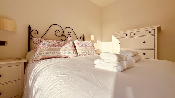 Una cama bien hecha con almohadillas estampadas con motivos florales, una mesita de noche con lámpara y un aparador con cajones.