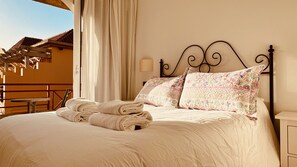 Un lit bien fait, avec des serviettes pliées, un coussin à motifs, une lampe fixée au mur et une vue sur un bâtiment dont le toit est recouvert de tuiles.