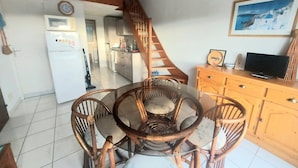 Un comedor con una mesa redonda y sillas, una cocina al fondo, una escalera y un televisor sobre un mueble de madera.