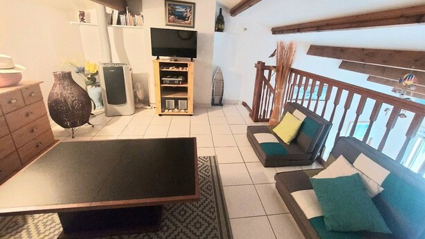 Una sala de estar con techo de madera, un televisor sobre un mueble de madera, un sofá con cojines, una mesa de comedor y una alfombra estampada.