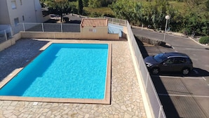 Una piscina con solárium de piedra, rodeada por una valla, y un estacionamiento con un coche.