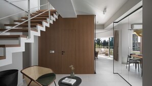 Ein modernes Interieur mit Treppe, einer Holztür, einem Korbstuhl, einem kleinen Tisch mit einem Buch und Blick ins Freie.