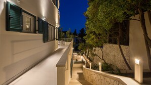 Ein Gebäude bei Nacht, mit einer Treppe, die zu einem Balkon führt, umgeben von Bäumen und einer Steinmauer.