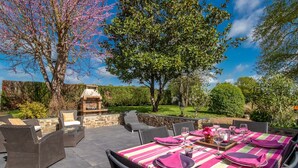 Un área de comedor al aire libre con una barbacoa de piedra, mobiliario de mimbre y vista a un jardín con árboles y seto.