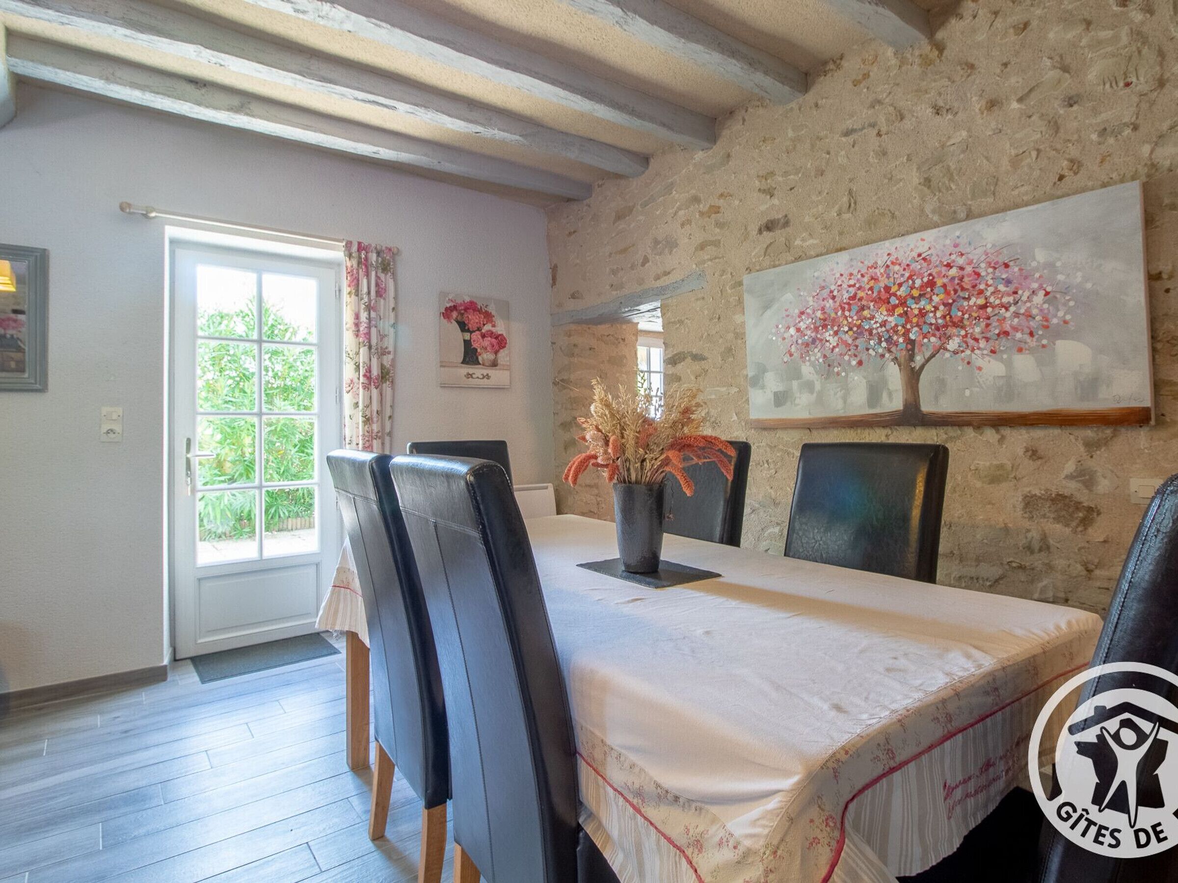 Un coin repas avec un mur en pierre, un grand tableau représentant un arbre et une fenêtre avec des rideaux.