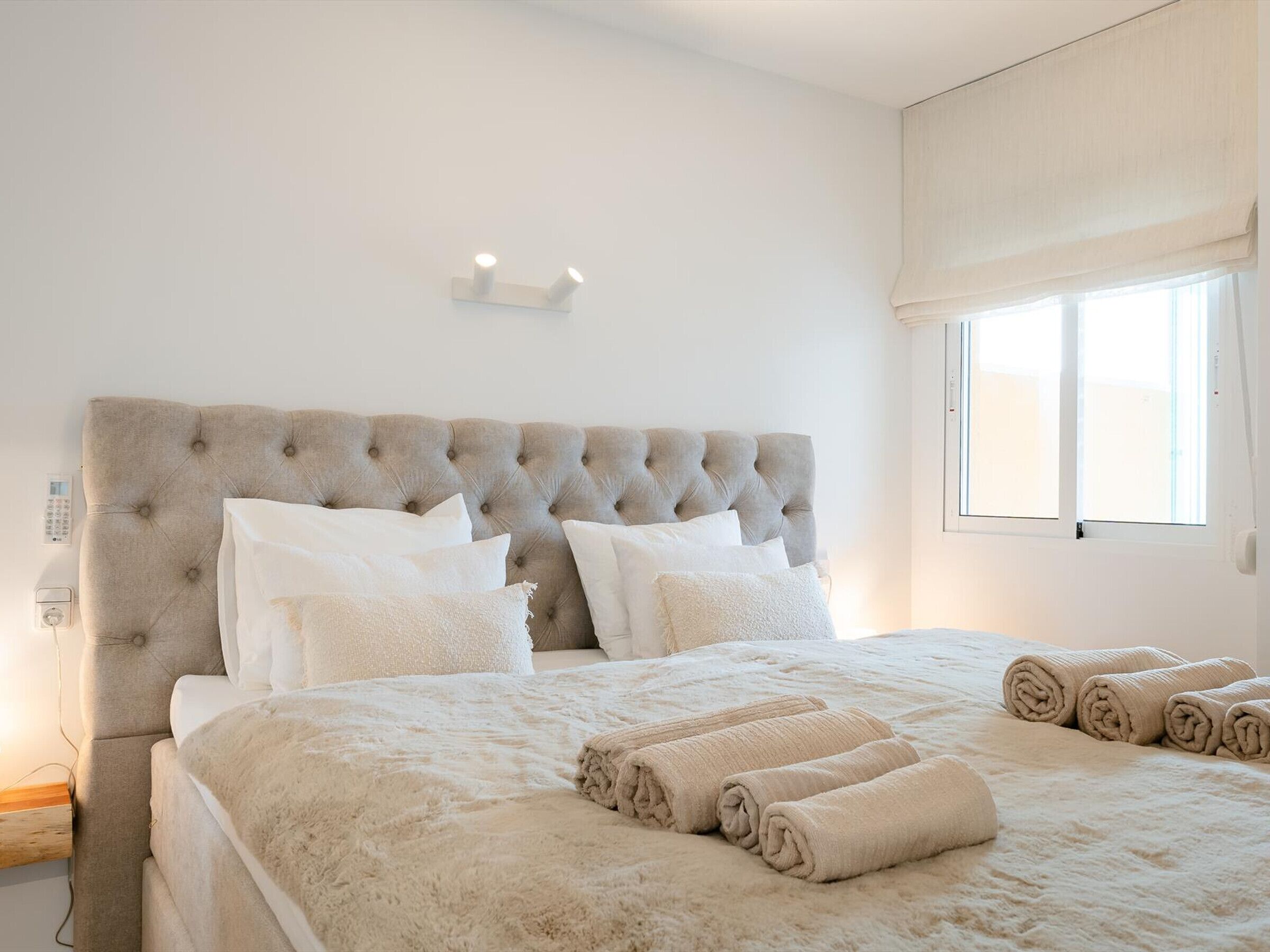 A bedroom with a tufted headboard, white bedding, and rolled towels on the bed.