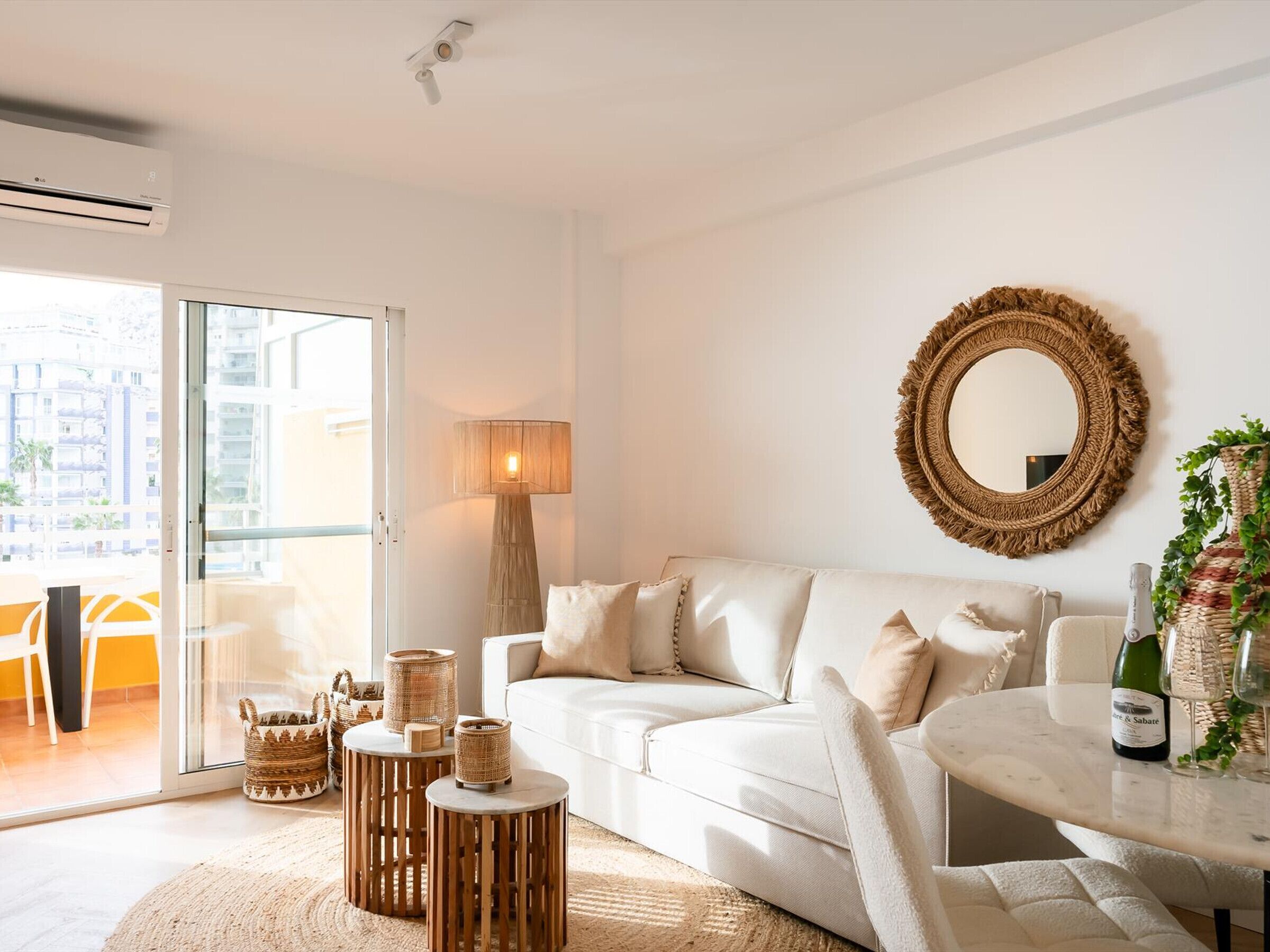 A modern living room with a white sofa, a round mirror, wicker baskets, and a table with a bottle and glasses.