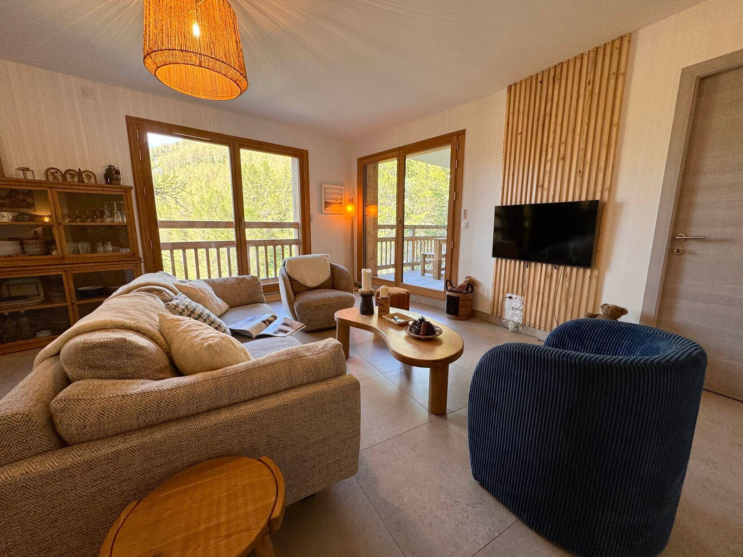 A spacious living room with a sofa, armchair, and ottoman, a wooden coffee table, and a television mounted on the wall.