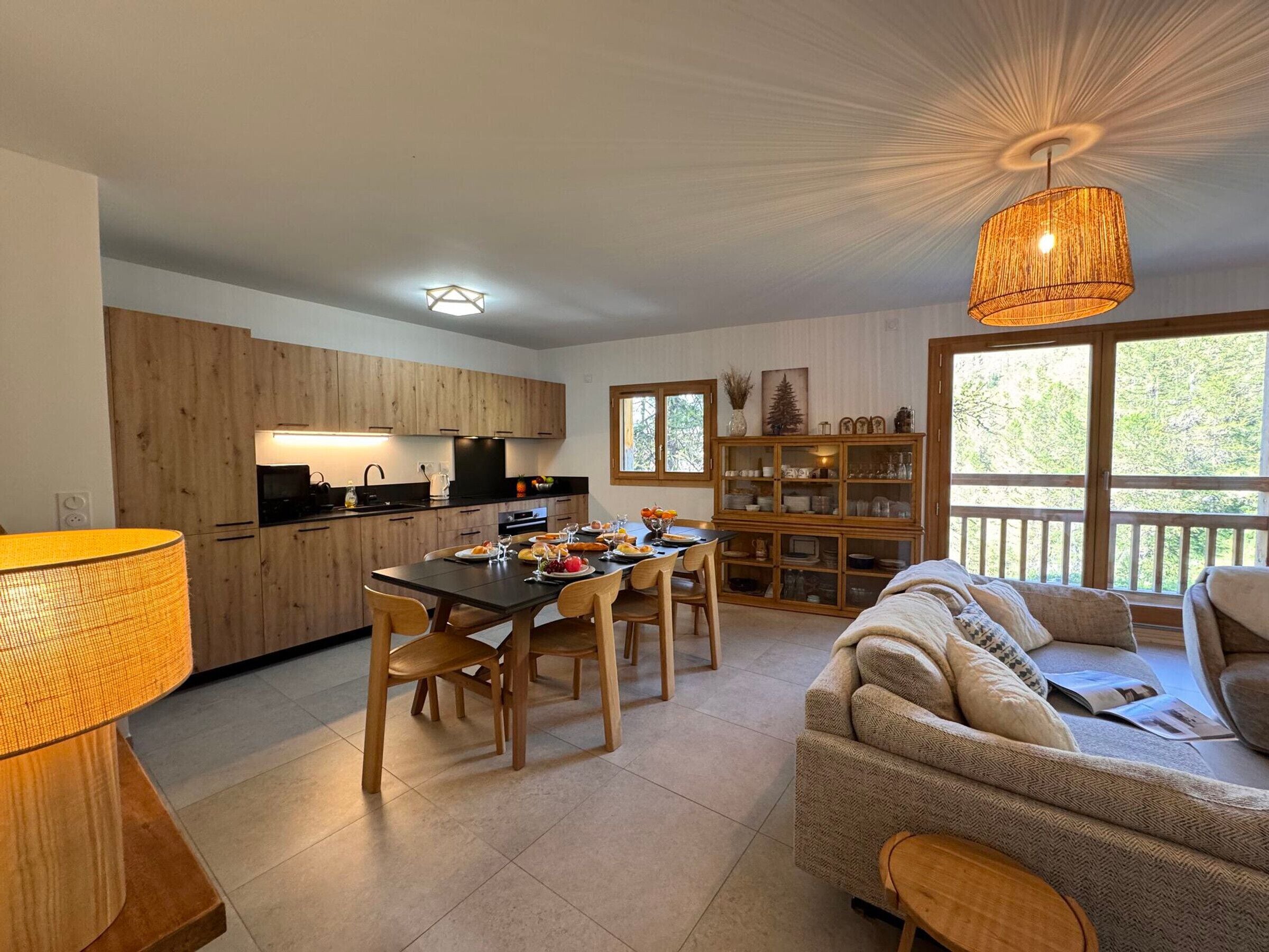 A modern kitchen with wooden cabinets, a dining table with chairs, a sofa, and a large window with a view of trees.