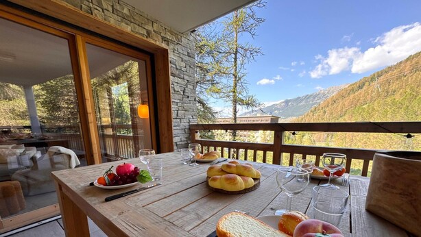 Een houten eettafel buiten, met uitzicht op bergen, een glas water, brood en fruit.