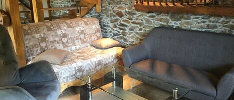 A cozy living area with a patterned sofa, a blue armchair, and a glass coffee table. A wooden staircase and stone wall are in the background.