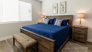 Quarto com cama de madeira, duas mesas de cabeceira com luminárias, um banquinho e janela com persianas.