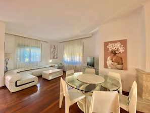 A modern living room with a glass dining table, white sofas, and a painting on the wall.