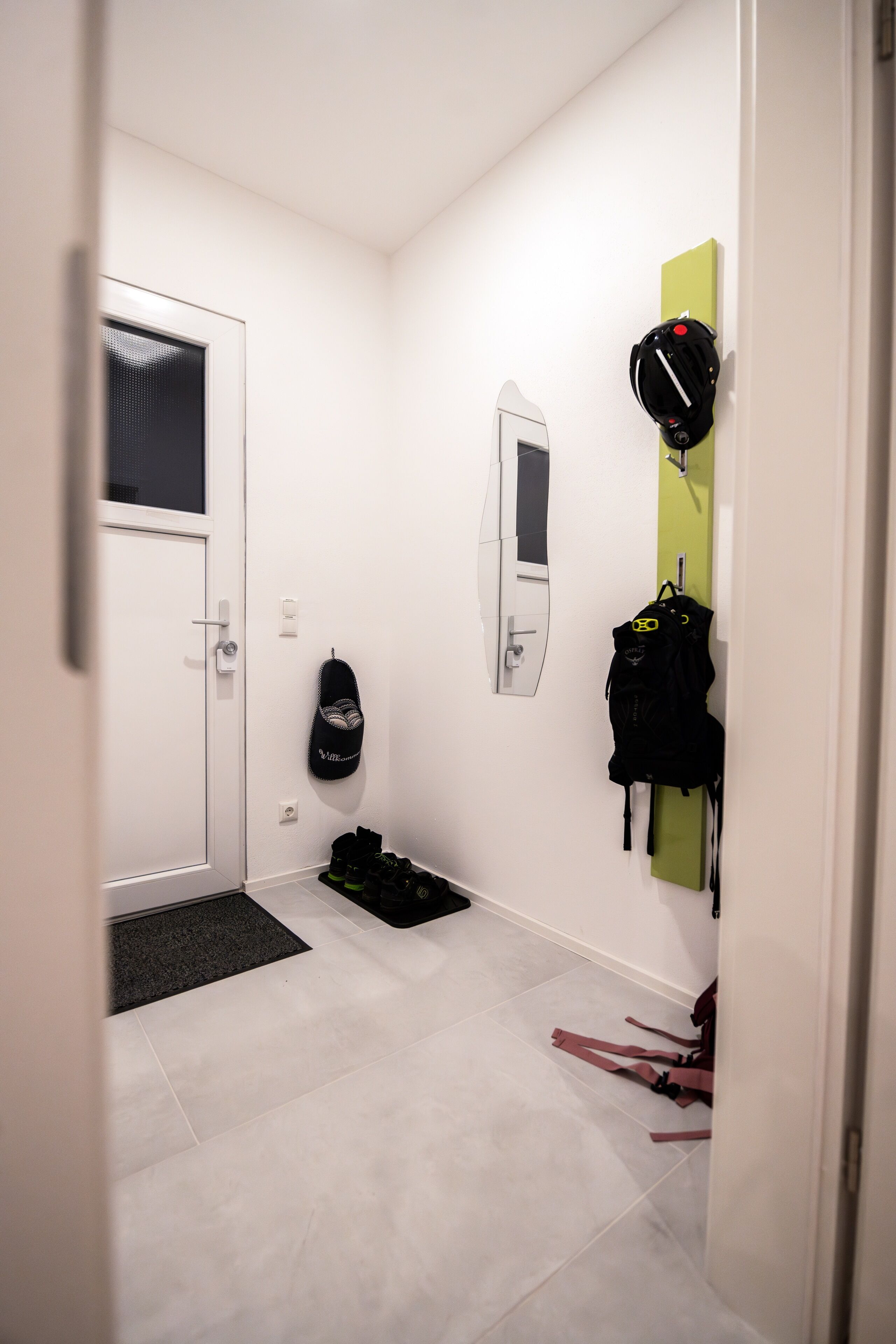 Um corredor com uma porta branca, um espelho e um suporte na parede com uma mochila preta e um capacete.