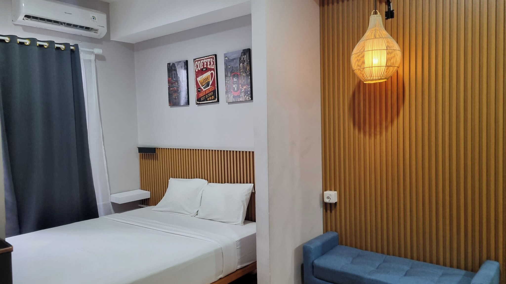 A hotel room with a bed, a nightstand, a blue chair, a wall-mounted lamp, and two posters on the wall.