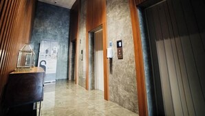 A hallway with a wooden panel wall, a marble floor, and an elevator with a control panel. - The Cozy Rooms at The Ayoma Residence (South Tangerang)