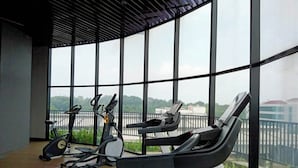 A gym with treadmills, elliptical machines, and stationary bikes, large windows with a view of greenery and buildings. - The Cozy Rooms at The Ayoma Residence (South Tangerang)
