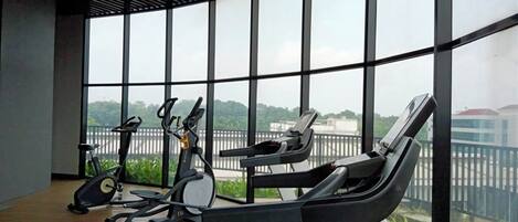 A gym with treadmills, elliptical machines, and stationary bikes, large windows with a view of greenery and buildings.
