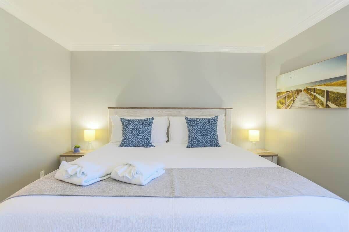 A neatly made bed with a white duvet, two patterned pillows, and a folded towel on a light-colored bedspread. There are two bedside lamps on either side of the bed.