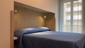 Ein Schlafzimmer mit Bett, Holz-Kopfteil und Fenster mit durchsichtigen Vorhängen.